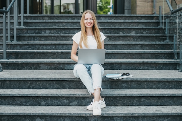 Chica joven estudiante sentada con un portátil en las escaleras cerca de la universidad sonriendo trabajando y mirando la cámara