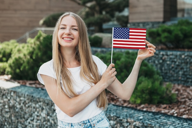 Chica joven estudiante sonriendo y muestra una pequeña bandera americana y se encuentra en el contexto de la universidad