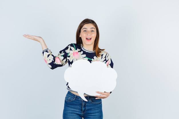 Chica joven extendiendo la palma en blusa floral, jeans y mirando alegre, vista frontal.