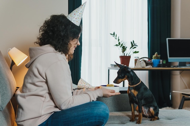 Chica joven feliz dando pastel casero a su perro, en el interior. Concepto de celebrar la fiesta de cumpleaños del perro.