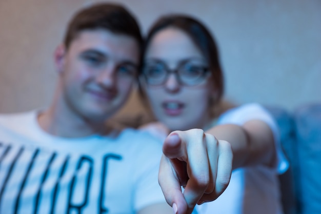 Chica joven con gafas está apuntando al foco de la cámara en su dedo sentado en un sofá con su novio en casa viendo la televisión, vista frontal en el resplandor azul del set