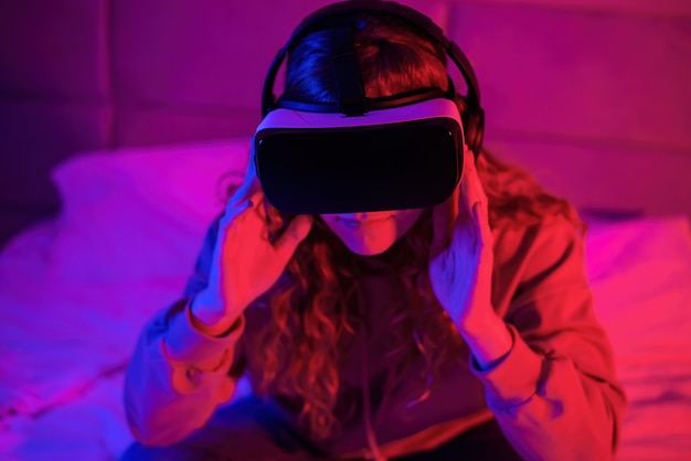 Chica joven en gafas de realidad virtual con iluminación azul y roja en la habitación En la cama. Entretenimiento en casa
