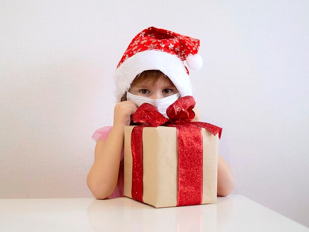 Chica joven con gorro de Papá Noel rojo y máscara de fondo blanco. Mascarilla quirúrgica bebé triste con regalo de Navidad. Concepto de Navidad Covid 19.