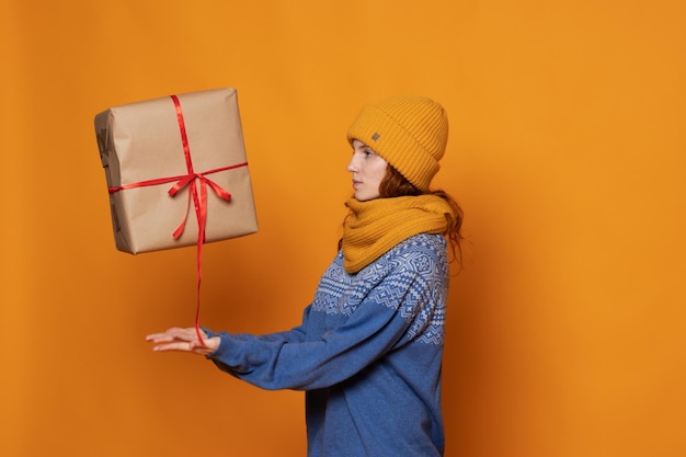 Chica joven con un gorro y un suéter sostiene un regalo sobre un fondo amarillo