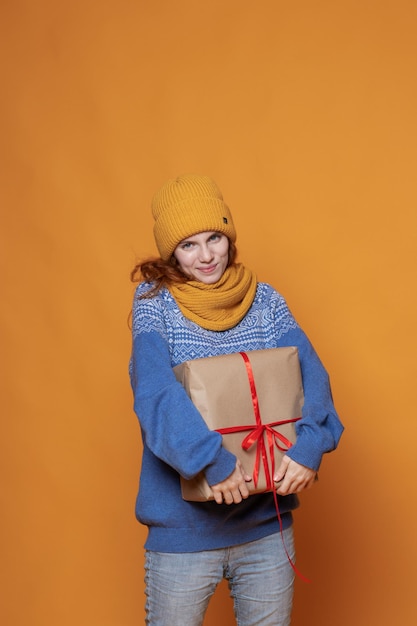 Chica joven con un gorro y un suéter sostiene un regalo sobre un fondo amarillo