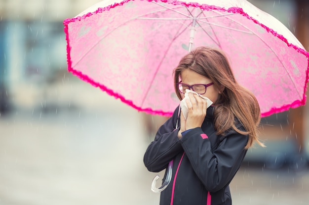 Chica joven con gripe sonándose la nariz con un pañuelo de papel bajo la lluvia de primavera