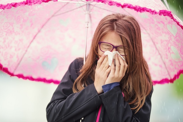 Chica joven con gripe sonándose la nariz con un pañuelo de papel bajo la lluvia de primavera