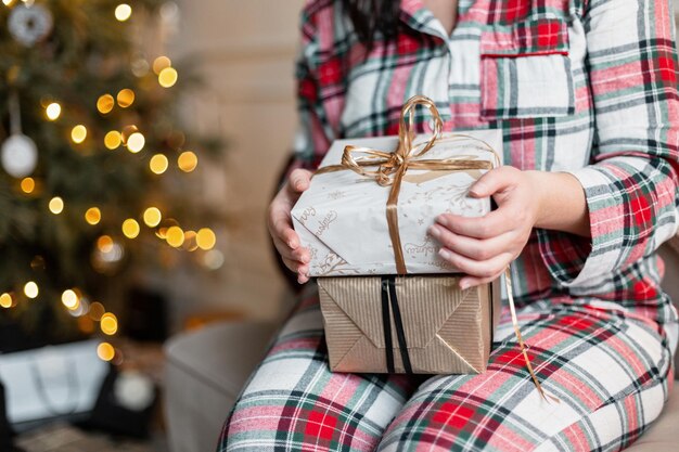 Una chica joven y guapa con pijamas a cuadros festivos se sienta en el sofá y sostiene regalos de Navidad cerca del árbol de Navidad con luces en vacaciones y regalos de Navidad