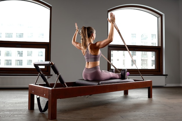 Chica joven haciendo ejercicios de pilates con una cama reformadora. Entrenador de fitness delgado hermoso sobre fondo gris reformador, bajo perfil, luz artística. Concepto de fitness.