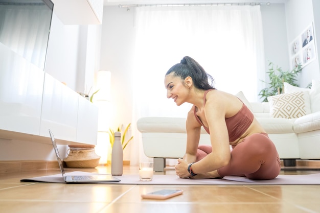 CHICA JOVEN HACIENDO YOGA Y PILATES EN EL SALÓN DE SU CASA CON LA COMPUTADORA