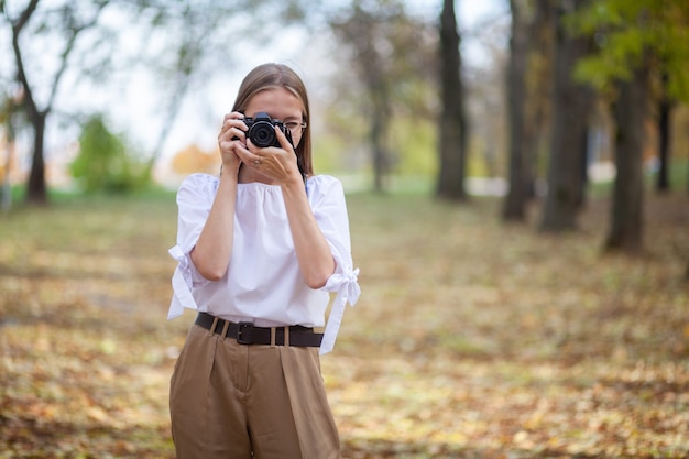 Foto chica joven hermosa atractiva que sostiene la cámara sin espejo moderna en el parque del otoño