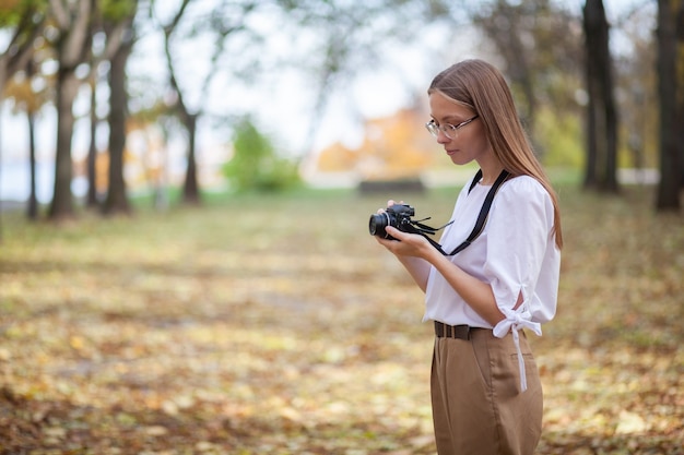 Foto chica joven hermosa atractiva que sostiene la cámara sin espejo moderna en el parque del otoño