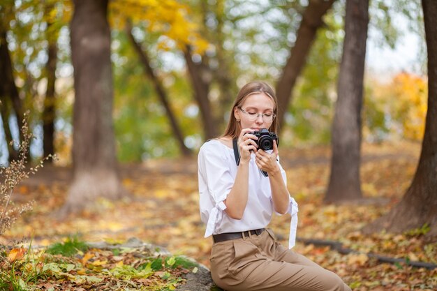 Foto chica joven hermosa atractiva que sostiene la cámara sin espejo moderna en el parque del otoño