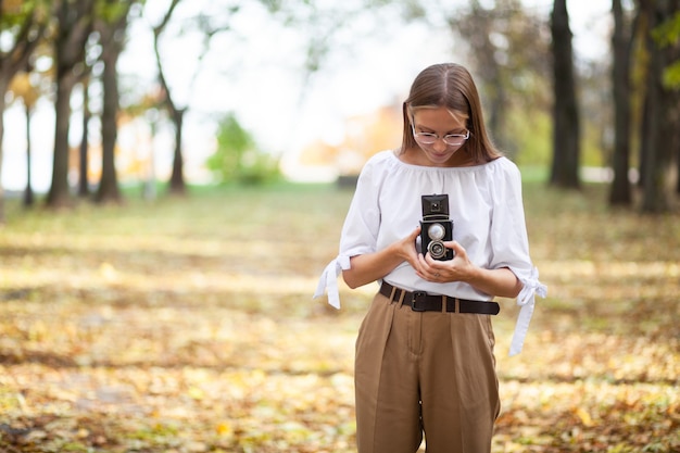 Foto chica joven hermosa atractiva que sostiene la cámara retro de la reflexión de la lente doble del vintage en el parque del otoño