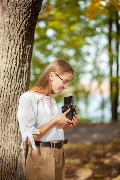 Foto chica joven hermosa atractiva que sostiene la cámara retro de la reflexión de la lente doble del vintage en el parque del otoño