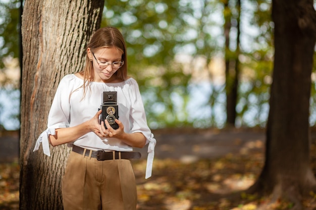 Foto chica joven hermosa atractiva que sostiene la cámara retro de la reflexión de la lente doble del vintage en el parque del otoño