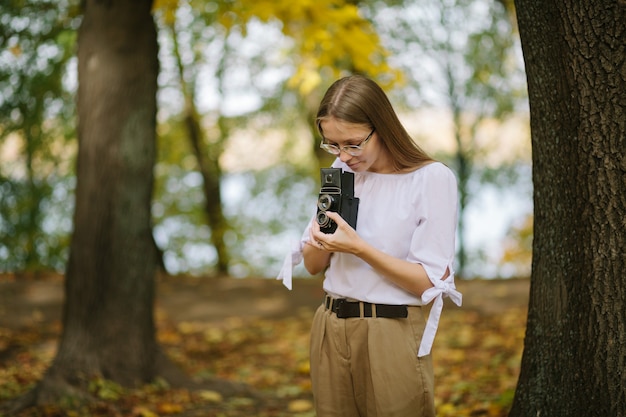 Foto chica joven hermosa atractiva que sostiene la cámara retro de la reflexión de la lente doble del vintage en el parque del otoño