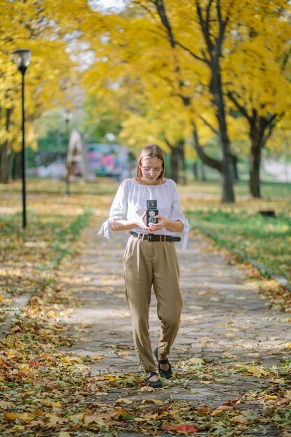 Chica joven hermosa atractiva que sostiene la cámara retro de la reflexión de la lente doble del vintage en el parque del otoño