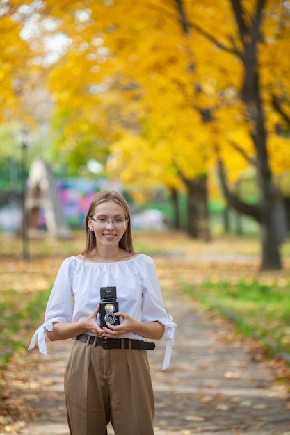 Chica joven hermosa atractiva que sostiene la cámara retro de la reflexión de la lente doble del vintage en el parque del otoño