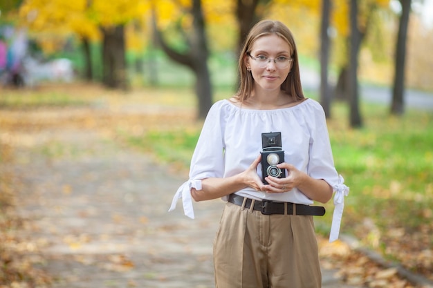 Foto chica joven hermosa atractiva que sostiene la cámara retro de la reflexión de la lente doble del vintage en el parque del otoño