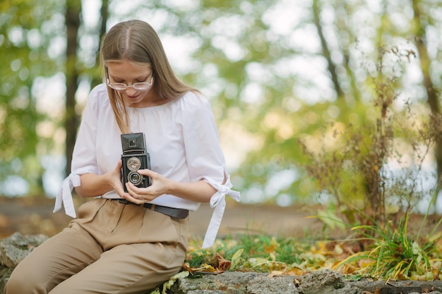 Chica joven hermosa atractiva que sostiene la cámara retro de la reflexión de la lente doble del vintage en el parque del otoño