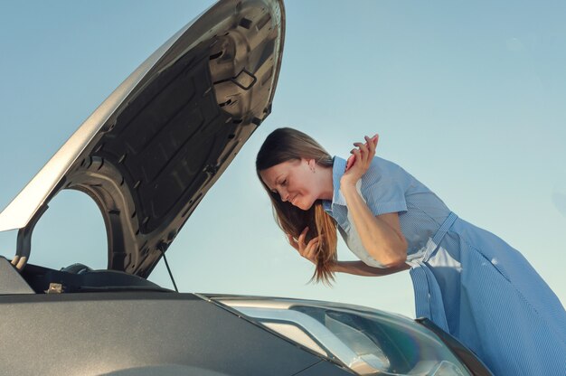 Chica joven y hermosa cerca de un coche roto con el capó abierto. problemas con el automóvil, no arranca, no funciona.