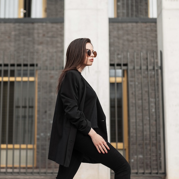 Chica joven hermosa elegante con gafas de sol en un blazer negro y pantalones camina en la calle