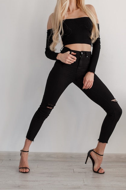 Chica joven hermosa elegante en ropa negra de moda con jeans negros y zapatos posando junto a la pared blanca