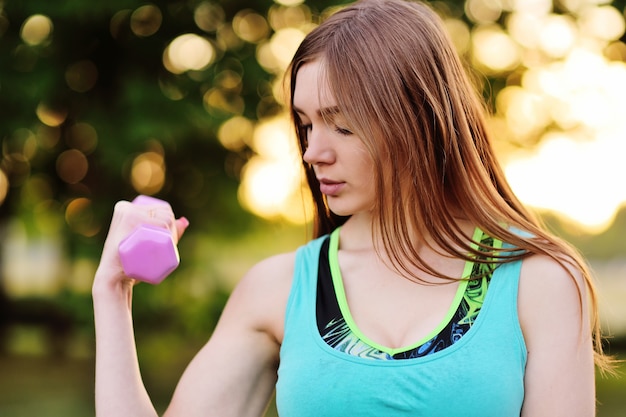 La chica joven hermosa está entrenando al músculo con el bíceps con pesas de gimnasia en el parque en un fondo de la naturaleza