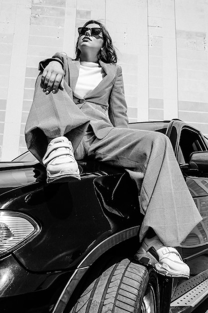 Una chica joven y hermosa está sentada en el capó de un automóvil. Una chica elegante con traje y gafas en un coche negro. Fotografía en blanco y negro.