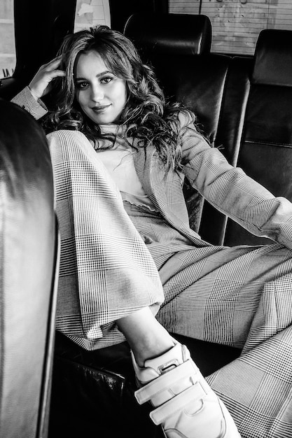 Una chica joven y hermosa está sentada en un coche. Chica con estilo en un gran retrato de traje, en el interior negro del coche. Fotografía en blanco y negro.