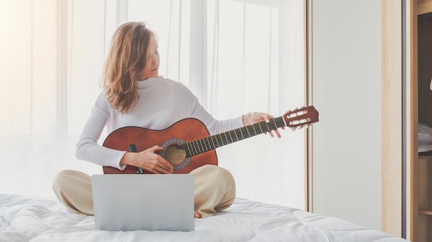 Chica joven hermosa que se sienta tocando la guitarra en cama en dormitorio