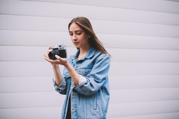 Chica joven hipster revisando fotos en la cámara retro mientras está de pie cerca de la pared blanca de moda