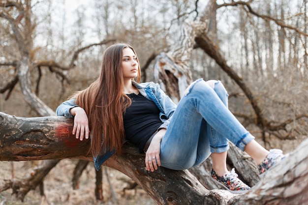 Chica joven inconformista con cabello largo y una chaqueta de mezclilla acostado en un árbol