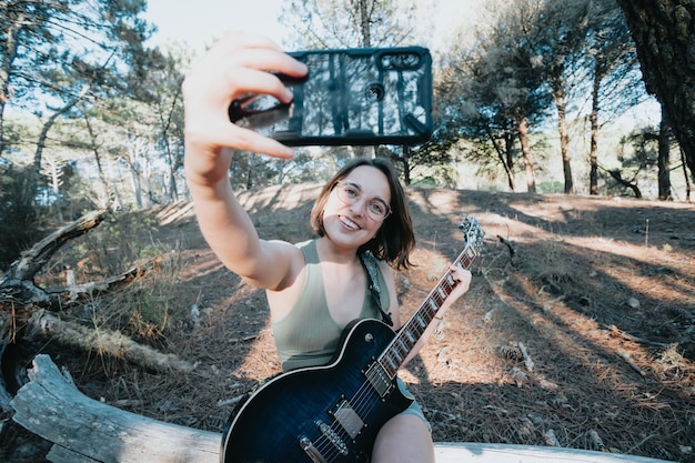 Chica joven inconformista con gafas tomando un selfie mientras sostiene una guitarra eléctrica, estudia conceptos musicales, copia espacio, redes sociales, superación personal, estilo de verano