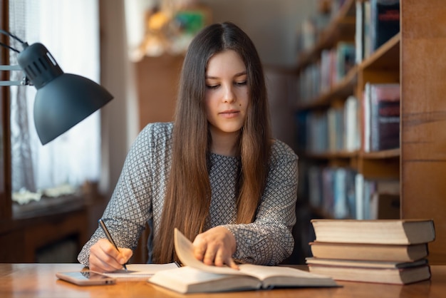 Chica joven inteligente leyendo un libro y tomando notas escribiendo la información más importante que lee Chica estudiante estudiando para los exámenes en la biblioteca universitaria trabajando en el escritorio