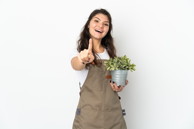 Chica joven jardinero ruso sosteniendo una planta aislada mostrando y levantando un dedo