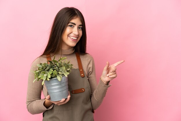 Chica joven jardinero sosteniendo una planta aislada en la pared rosa apuntando hacia atrás