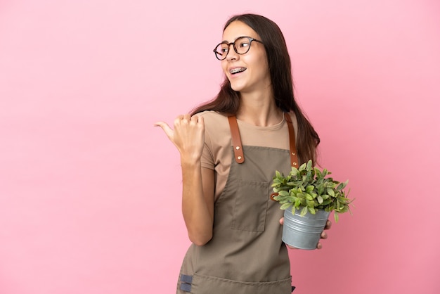 Chica joven jardinero sosteniendo una planta aislada sobre fondo rosa apuntando hacia el lado para presentar un producto