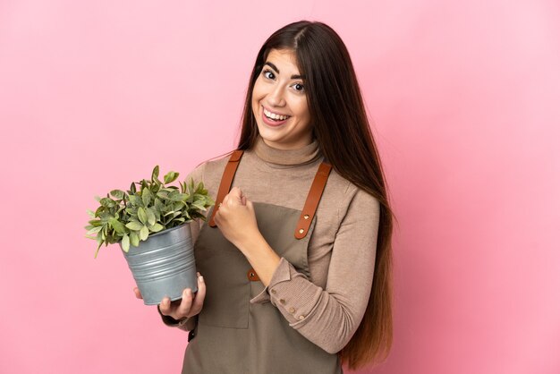 Chica joven jardinero sosteniendo una planta aislada sobre fondo rosa celebrando una victoria