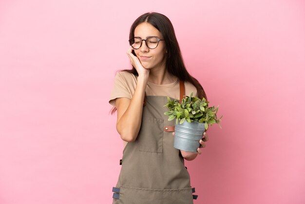 Chica joven jardinero sosteniendo una planta aislada sobre fondo rosa frustrado y cubriendo las orejas