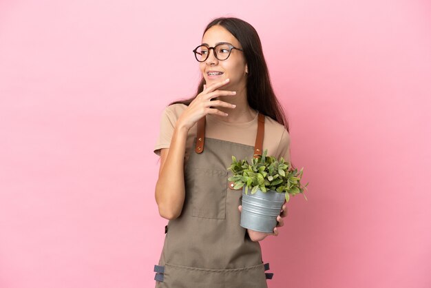 Chica joven jardinero sosteniendo una planta aislada sobre fondo rosa mirando hacia arriba mientras sonríe