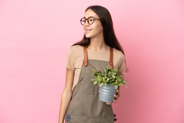 Chica joven jardinero sosteniendo una planta aislada sobre fondo rosa mirando hacia el lado y sonriendo