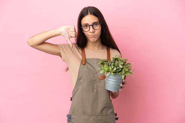 Chica joven jardinero sosteniendo una planta aislada sobre fondo rosa mostrando el pulgar hacia abajo con expresión negativa