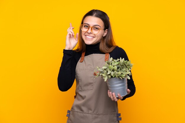 Chica joven jardinero sosteniendo una planta sobre amarillo aislado con cruzar los dedos