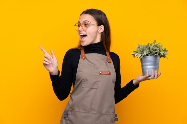 Chica joven jardinero sosteniendo una planta sobre dedo acusador amarillo aislado al lado