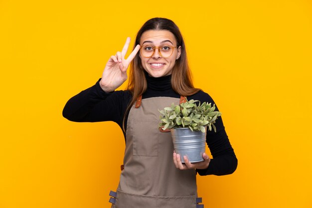 Chica joven jardinero sosteniendo una planta sonriendo y mostrando el signo de la victoria