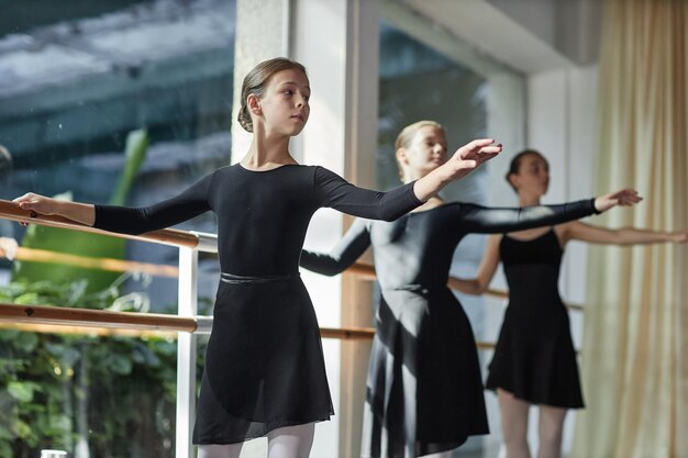 Chica joven en leotardo negro levantando la mano de pie en la barra durante la clase de ballet en el estudio de danza