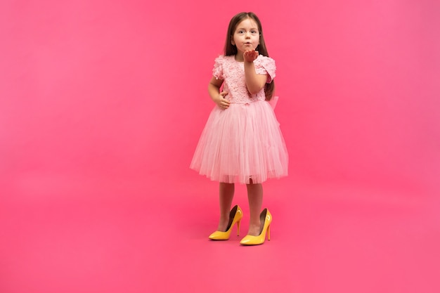 Chica joven linda de moda con cabello largo morena, en elegante vestido con zapatos de madre grande, enviando beso a la cámara aislada sobre fondo rosa. Divirtiéndose del niño alegre. Lugar para el texto.