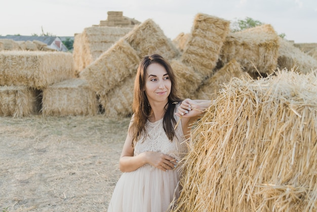 La chica joven lleva el vestido blanco del verano cerca de la bala de heno en campo.